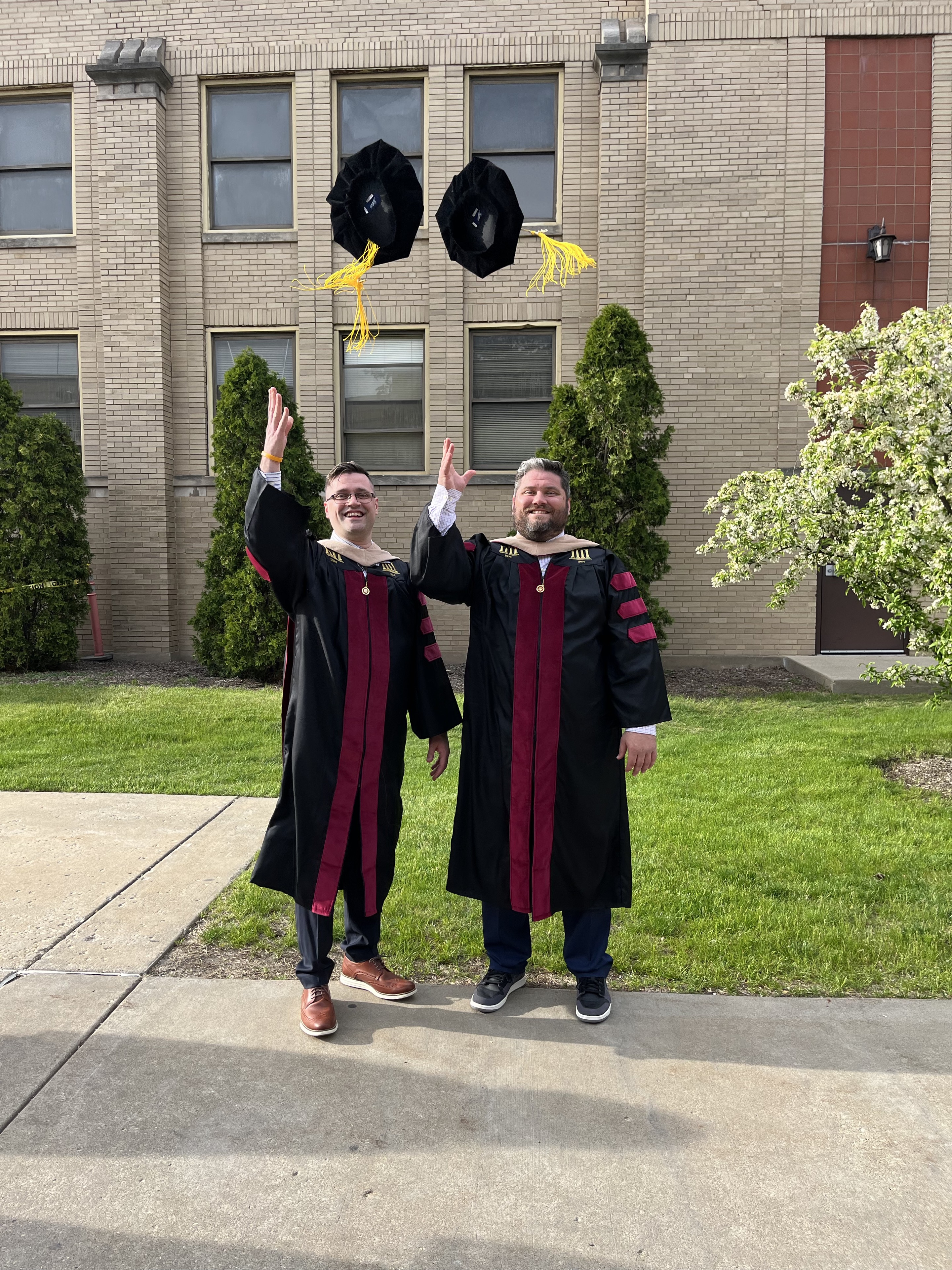 Two doctoral graduates celebrating outdoors, tossing their caps into the air